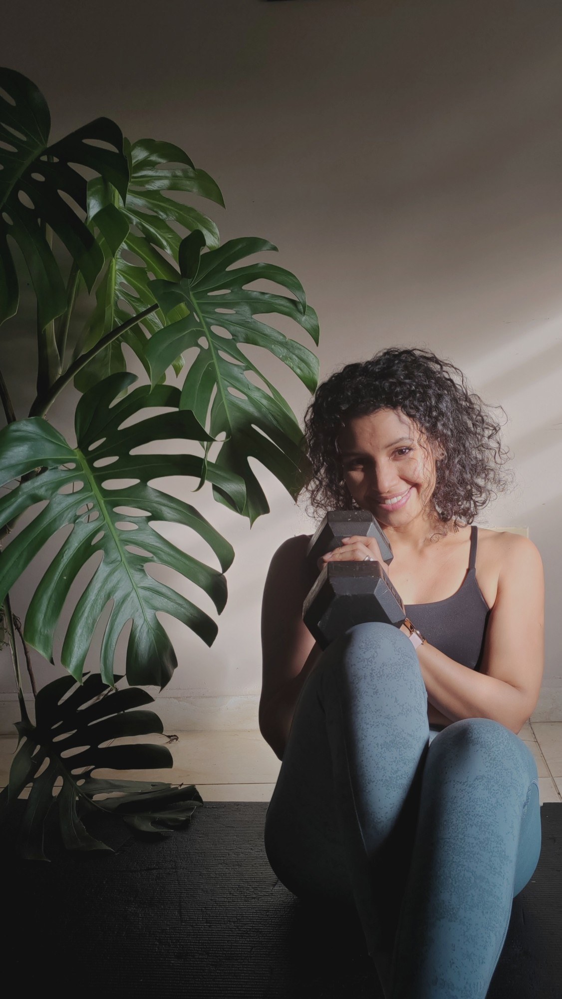 Leysi sentada en el piso de su casa, sonriendo, con una mancuerna apoyada
             en el hombro.